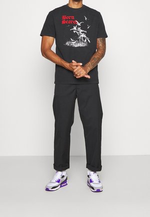 T-shirt noir avec un imprimé graphique en texte blanc et rouge, associé à un pantalon noir et des baskets multicolores. Tenue décontractée.