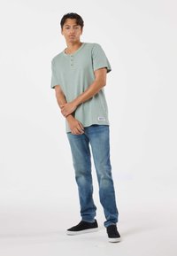 Ljusgrön henley-topp med korta ärmar, tre knappar och en sidolapp, tillsammans med blå jeans och svarta sneakers. Slät textur.