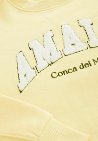 Gelbes Sweatshirt mit "AMAI" in weißer, strukturierter Stickerei und schwarzem Rand. "Conca dei Marmi" in schwarzer Schrift darunter gedruckt. Rippenkragen und -manschetten.