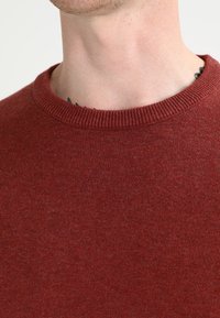 Bordový pletený svetr s kulatým výstřihem, který se vyznačuje jemnou texturou a žebrovanými detaily na límci. Materiál je hladký a rovnoměrný.