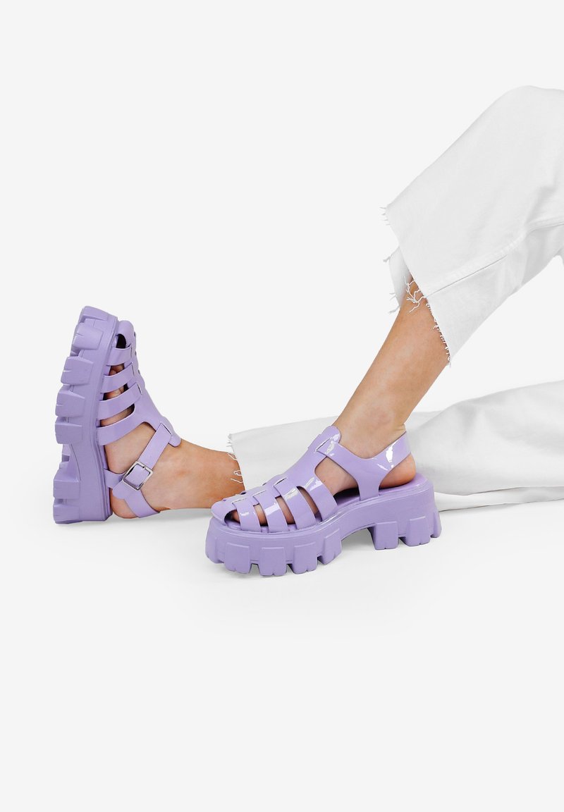 Zuecos de goma morados con la punta abierta y correas entrelazadas, que presentan una suela gruesa y texturizada, además de un detalle de hebilla.