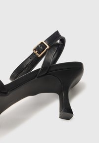 Schwarze Leder-Pumps mit hohen Absätzen, schmalem Knöchelriemen und goldfarbener Schnalle. Mit runder Zehenpartie und strukturiertem Schuhboden.