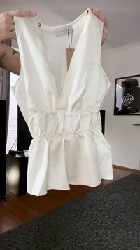 White sleeveless blouse with deep V-neck and ruched waist held up by tattooed hands indoors.
