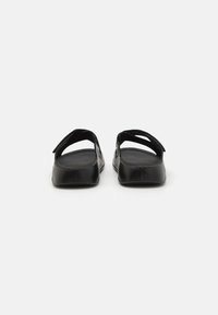 Scarpe nere piatte con retro aperto, realizzate in pelle liscia. Presentano cinturini regolabili e una suola piatta con dettagli minimi.