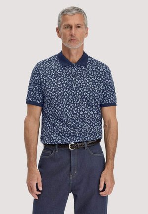Mittelalter Mann trägt ein marineblaues Poloshirt mit weißen Blumenmustern, dunkelblaue Jeans und einen schwarzen gewebten Gürtel, steht vor einem einfarbigen Hintergrund.