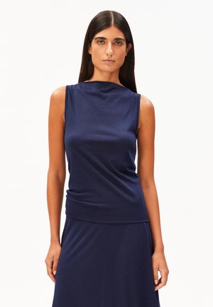 Femme aux longs cheveux foncés portant un haut sans manches bleu marine et une jupe assortie, debout devant un fond blanc uni.