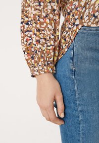Brun, mønsteret langærmet bluse med farverige pletter og puffede ærmer, parret med lyseblå denim jeans. Glat tekstur.