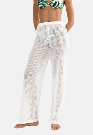 Kobieta ubrana w białe plażowe spodnie z siateczki ze sznurkiem w pasie, stojąca boso na jednolitym tle.