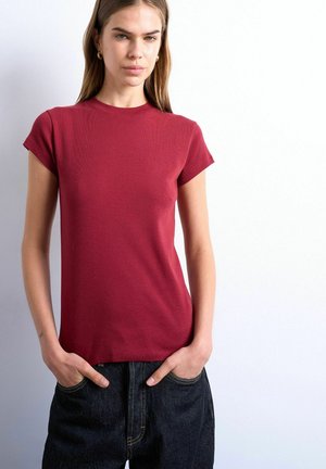 Young woman with long hair wearing a fitted red short-sleeve top and dark loose jeans with hands in pockets against a white background.