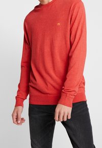 Pull rouge à manches longues avec col rond en tissu jersey doux, présentant un logo discret jaune sur la poitrine et des poignets et ourlet côtelés.