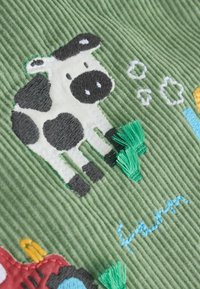 Le tissu en velours côtelé brodé présente une vache noire et blanche, de l'herbe verte et des éléments colorés, y compris des accents bleus et jaunes sur un fond vert atténué.