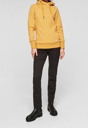 Personne portant un sweat à capuche jaune moutarde, un pantalon noir slim et des bottines noires, debout devant un fond clair uni.