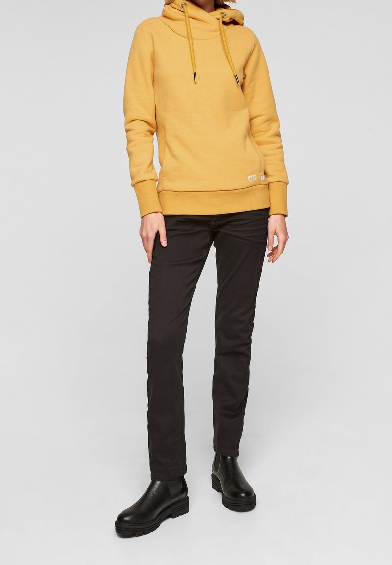 Personne portant un sweat à capuche jaune moutarde, un pantalon noir slim et des bottines noires, debout devant un fond clair uni.