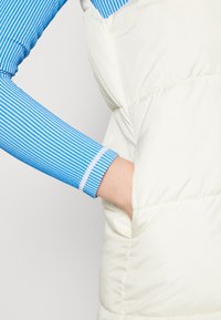 White padded vest with a side pocket, paired with a blue and white striped long-sleeve shirt, showing arm detail and fabric texture.
