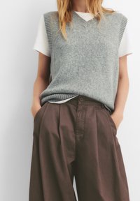 Grijze gebreide vest met een V-hals over een wit T-shirt, gecombineerd met losse bruine broeken met plooien en zijzakken.
