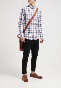 Man in zwarte broek, bruine schoenen, wit geruit shirt en met een bruine leren schoudertas, staande op een witte achtergrond.