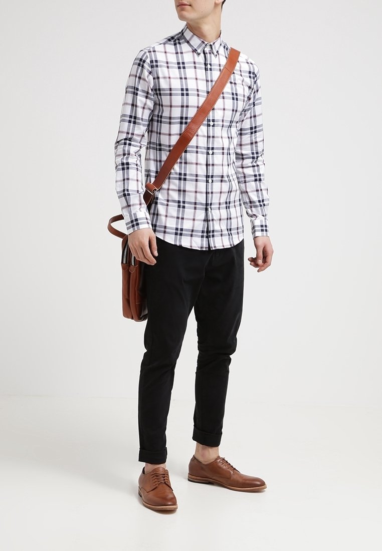 Man in zwarte broek, bruine schoenen, wit geruit shirt en met een bruine leren schoudertas, staande op een witte achtergrond.
