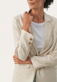 Beige lina blazer dengan desain yang disesuaikan, kerah notch, dan dua aksen tombol di lengan. Dipadukan dengan atasan putih bergaris.