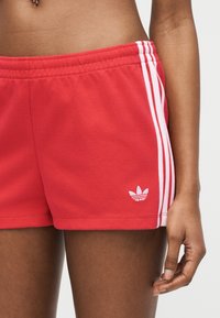 Shorts de sport rouges avec des bandes blanches sur les côtés et un petit logo tréfle, portés par une personne à la peau foncée avec le bras détendu le long du corps.