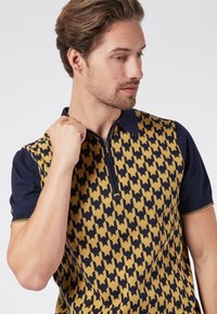 Polo shirt featuring a navy blue collar and short sleeves, with a bold yellow houndstooth pattern on the body and a front zipper detail.
