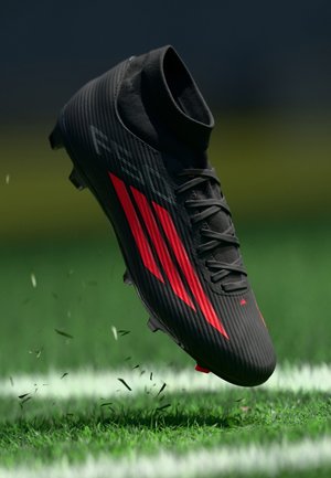 Schwarzer Fußballschuh mit roten Akzenten, strukturierter Obermaterial, hohem Knöcheldesign und Stollen für besseren Halt, über grünem Gras positioniert.