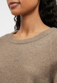Gros plan d'une personne portant un pull doux marron clair à col rond avec un tissu en maille texturée et de longs cheveux bouclés foncés visibles.