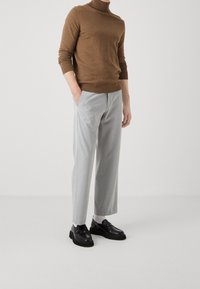 Maglione a collo alto marrone realizzato in materiale morbido, abbinato a pantaloni grigi chiari plissettati. Mocassini neri completano l'outfit.