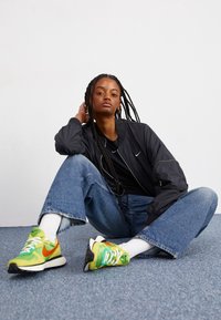 Svart bomberjacka, svart T-shirt, ljusblå oversize jeans och färgglada Nike-sneakers i grönt, orange och vitt, med vita strumpor.