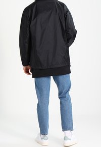 Svart nylonbomberjacka med ribbad fåll, kombinerad med ljusblå denimjeans och vita sneakers, sedd bakifrån.