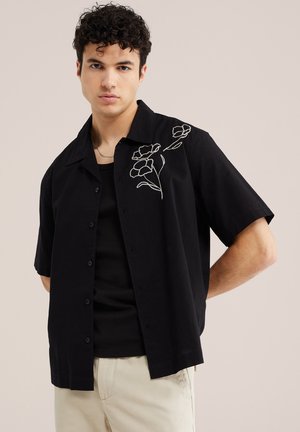 Jeune homme portant une chemise noire à manches courtes avec une broderie florale blanche sur la poitrine, un pantalon beige et un collier en chaîne.