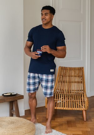 Navy t-shirt, plaid shorts in blue, white, and red. Standing on hardwood floor with woven stool and rattan chair in background.