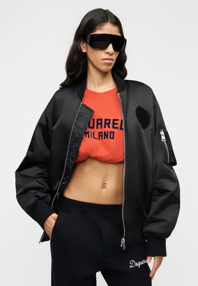 Μαύρο bomber μπουφάν με φερμουάρ μπροστά, φαρδιά εφαρμογή και μανσέτες στα μανίκια· κόκκινο t-shirt με το κείμενο "SQUARED MILANO"· μαύρη φόρμα με λογότυπο.
