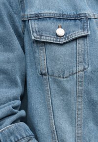 Κοντινό πλάνο τσέπης ανοιχτό μπλε τζιν μπουφάν με ορατές ραφές και ασημένιο κουμπί στερεωμένο στην κλείστρα της τσέπης.