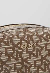 Ruskeakuvioinen DKNY-laukku, jossa on teksturoitu pinta, kullanvärinen logokoriste ja vetoketjusuljenta yläosassa.