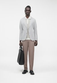 Blazer grigio chiaro, maglione beige e pantaloni marroni. Borsa tote nera, scarpe nere. Modella in piedi davanti a uno sfondo bianco neutro.