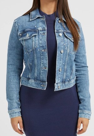 Kobieta w jasnoniebieskiej kurtce dżinsowej ozdobionej perłami, ubrana na granatową prążkowaną sukienkę, stojąca na tle o gładkim tle.