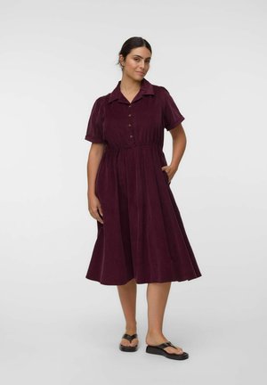 Vrouw staat en draagt een donkerpaarse midi-jurk met korte mouwen, knopen, kraag en zwarte platformsandalen, hand in de zijzak van de jurk.