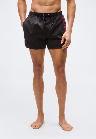 Homme torse nu portant un short de bain noir avec des poches latérales et un texte rouge sur la cuisse gauche, debout pieds nus sur un fond uni.