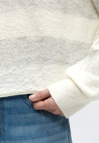 Hånd med neutral neglelak delvist i forlommen på blå jeans, iført blød hvid og lysegrå striped strikket sweater.