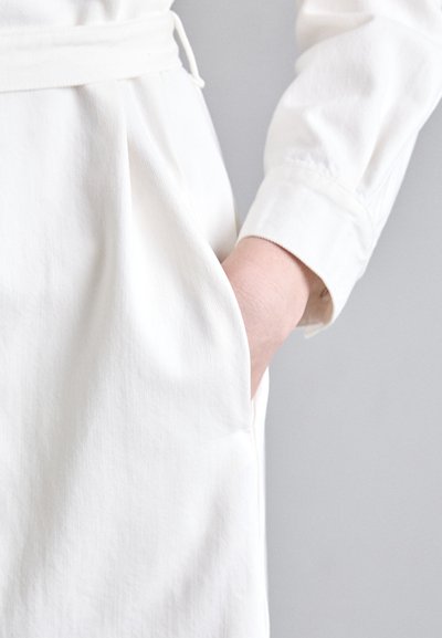 Balta medvilnės suknelė su ilgomis rankovėmis, turinti šoninę kišenę ir surišamą juosmens detalę. Lygi tekstūra ir paprastas, tvarkingas dizainas.