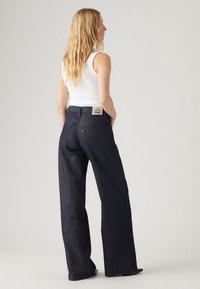 Σκούρες τζην παντελόνες σε φαρδιά γραμμή με ορατές πίσω τσέπες, με ένα λογότυπο στη μέση και λεία υφή. Φοριούνται με ένα εφαρμοστό άσπρο μπλουζάκι.