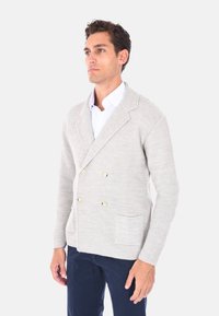 Cardigan doppio petto grigio chiaro con tessuto lavorato a maglia, bottoni color oro e due tasche frontali. Indossato sopra una camicia bianca.