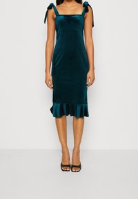 Teal sammetklänning med axelband, figurnära liv och volangkant. Slät yta med en lätt glans och minimalistisk design.