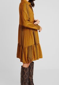 Langärmliges, senfgelbes Kleid mit vertikalen Streifen und floralen Akzenten, einem gestuften Rock und einem geknöpften Kragen. Kombiniert mit braunen Stiefeln im Schlangenledermuster.