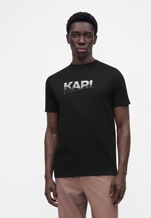 Jeune homme aux cheveux courts et bouclés portant un t-shirt noir avec le logo "KARL" et un pantalon brun clair, se tenant devant un fond blanc uni.