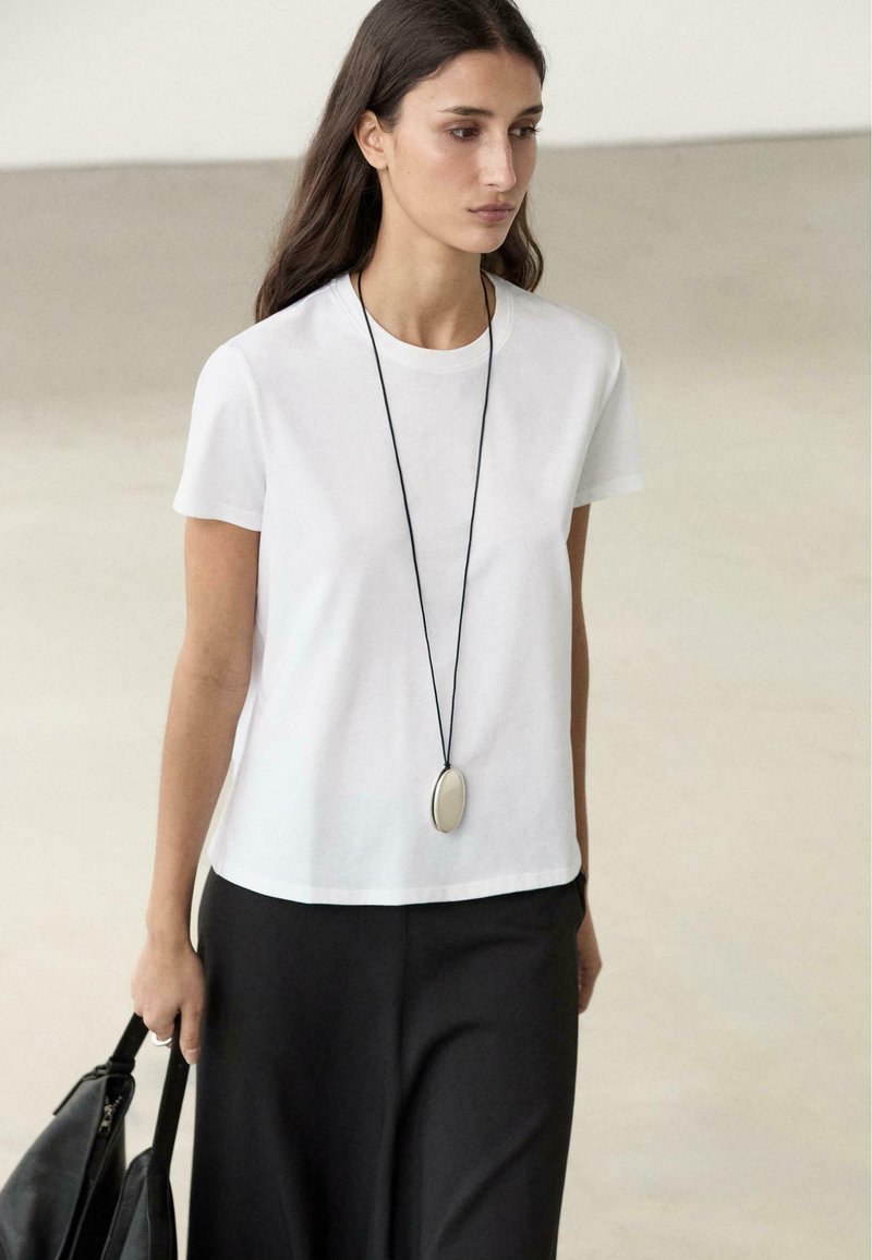 Frau trägt ein weißes T-Shirt, schwarzen Rock, lange ovale Anhänger-Halskette, hält eine schwarze Tasche vor neutralem Hintergrund.