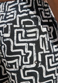 Pantalones cortos negros con una tela texturizada que presenta un patrón abstracto similar a un laberinto en blanco. Incluyen un bolsillo lateral con un botón de presión negro.