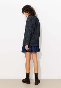 Veste tricotée texturée bleu marine, superposée à une jupe en denim évasée, portée avec des bottines brunes et des chaussettes gris foncé, vue de l'arrière.