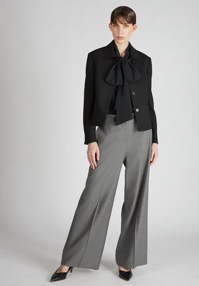 Blazer nero con colletto a grande fiocco, pantaloni grigi a gamba larga in un tessuto testurizzato e décolleté nere a punta, su uno sfondo neutro.