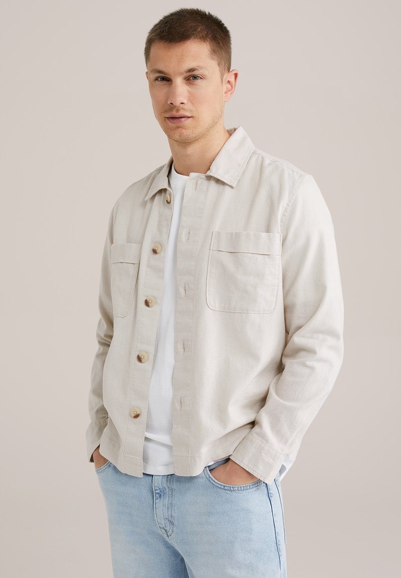 Licht beige overhemd met knopen, gemaakt van gestructureerde stof, met twee borstzakken en een klassieke kraag, gecombineerd met een wit T-shirt eronder.